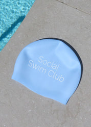 Blue swim cap with 'Social Swim Club' text on a concrete surface near a pool.
