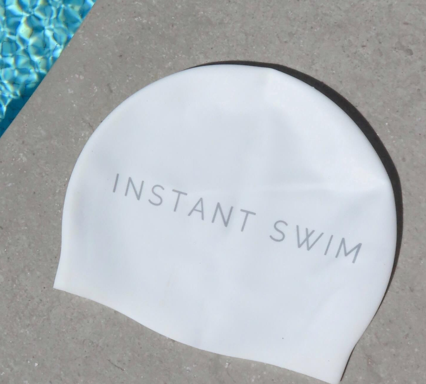 White swim cap with 'Instant Swim' text on a concrete surface next to a pool.