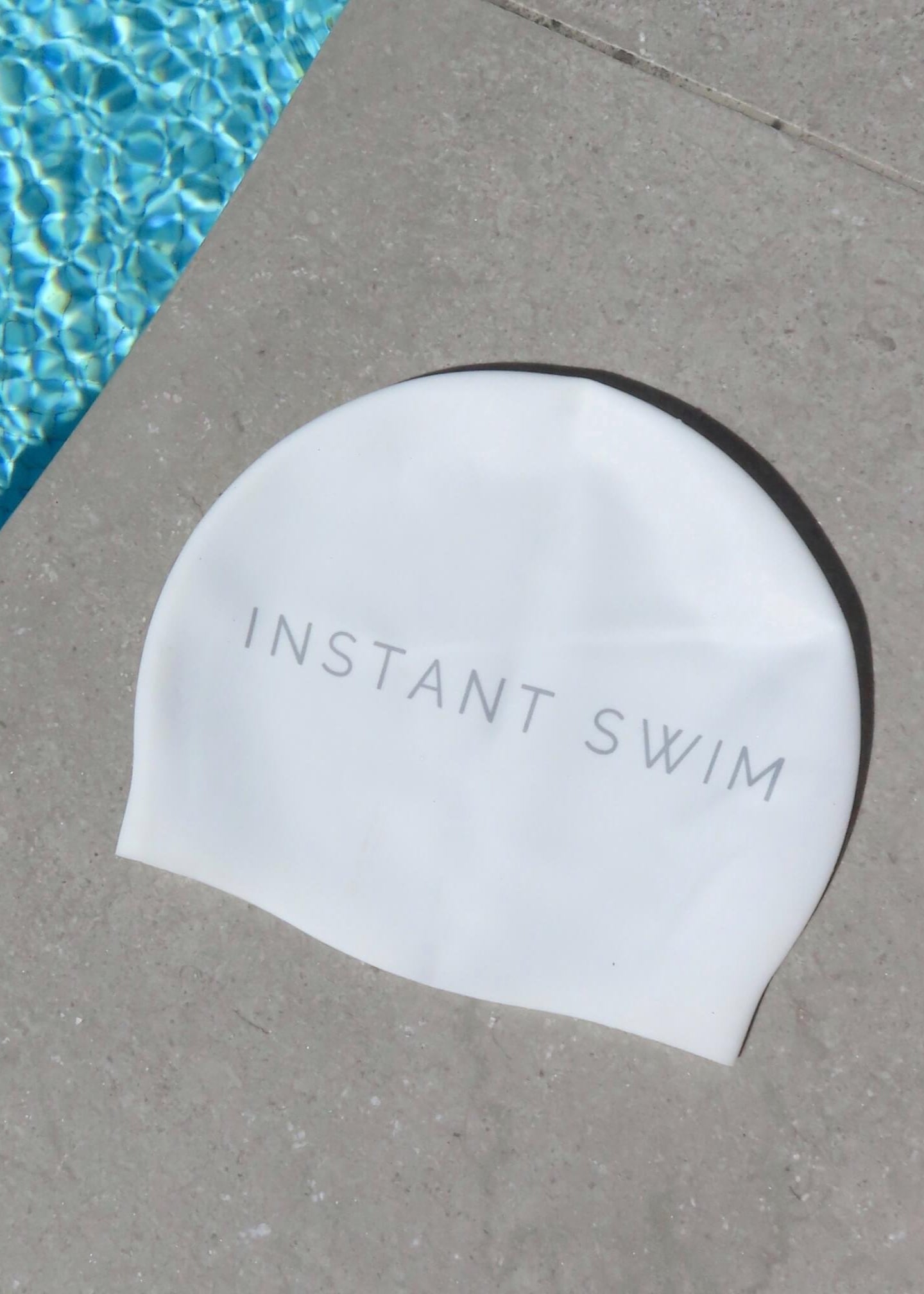 White swim cap with 'Instant Swim' text on a concrete surface next to a pool.