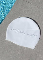 White swim cap with 'Instant Swim' text on a concrete surface next to a pool.